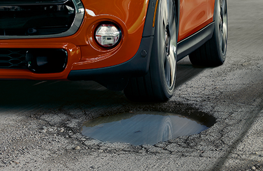 A close-up of the front of an orange MINI Cooper Hardtop about to hit a large pothole on an asphalt road.