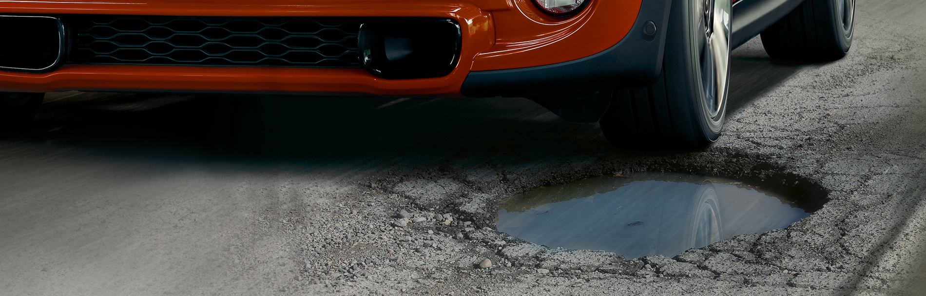 An extreme close-up of the front of an orange MINI Cooper Hardtop about to hit a large pothole on an asphalt road.
