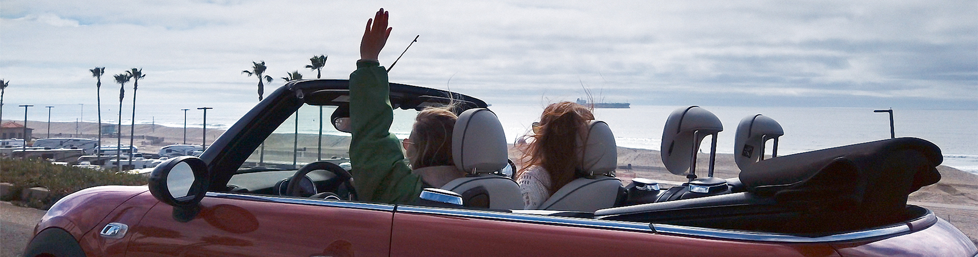 Female driver raising arm inside of a red MINI convertible with roof down near a beach.