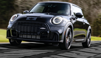 Black MINI John Cooper Works Hardtop riding on road made to look like a tire tread. Green trees and grass are in the background.