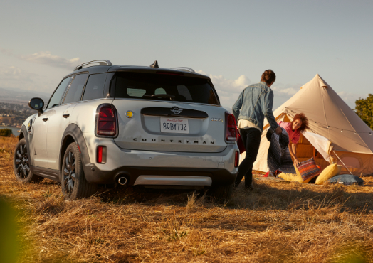 Silver MINI Countryman parked in a field. A man and a woman can be seen to the right of the car. There is a tent set up behind them.